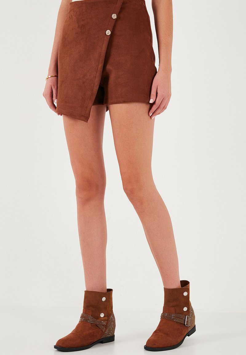 Shorts en daim brun à enrouler avec trois boutons, présentant une texture lisse. Associés à des bottines brunes à la cheville, ornées de clous et de sangles.