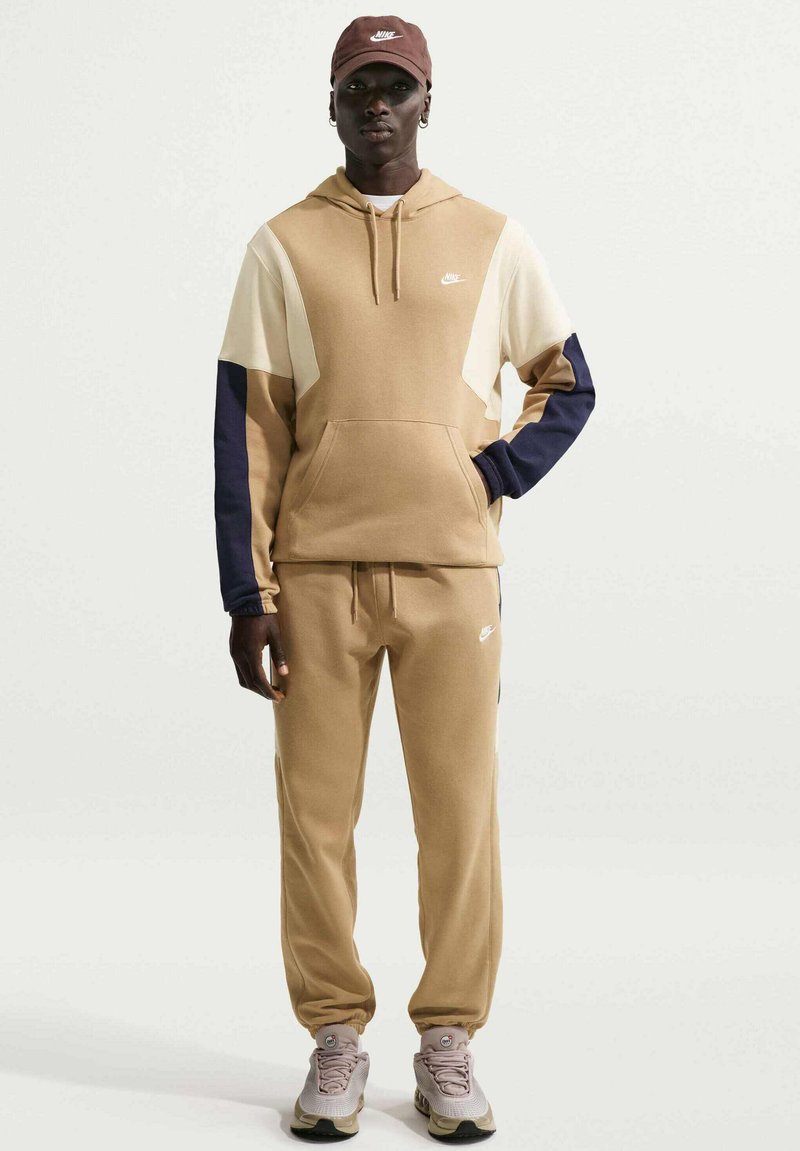 Chándal Nike beige y azul marino con sudadera con capucha y pantalones ajustados; presenta colores sólidos y un diseño elegante, con zapatillas a juego.