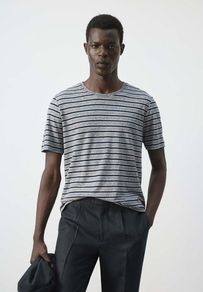 Jeune homme portant un t-shirt rayé gris et noir et un pantalon foncé tenant une veste sombre devant un fond clair uni.