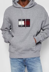 Sudadera gris con bolsillo tipo canguro y capucha con cordón. Presenta un parche de logo multicolor con texto en el pecho. Tejido de peso medio.