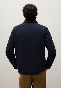 Hombre con cabello rizado que lleva una camisa de manga larga de color azul marino oscuro y pantalones marrones, visto desde atrás sobre un fondo claro y liso.
