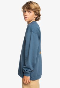 Sudadera azul con un corte relajado, puños y bajo acanalados. Presenta un detalle del logo en naranja en la parte posterior y una textura de algodón suave.