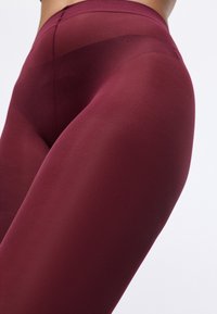 Bordeauxfarbene Leggings aus glattem, dehnbarem Material, mit hohem Bund und dezentem Glanz, die eine figurbetonte Silhouette und nahtloses Design präsentieren.