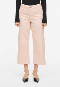 Femme portant un pantalon ample rose pâle coupé court, un haut noir à manches longues et des escarpins noirs à bout pointu, debout contre un fond uni.