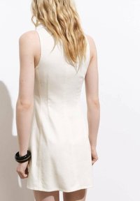 Robe sans manches de couleur crème avec une coupe ajustée, texture lisse et détails de couture subtils. Un bracelet noir orne le poignet du mannequin.