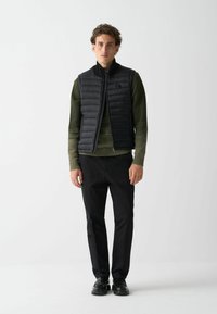 Svart vadderad väst med hög krage, bärs över en mörkgrön stickad tröja. Svarta byxor och skor kompletterar outfiten, med en sömlös design.