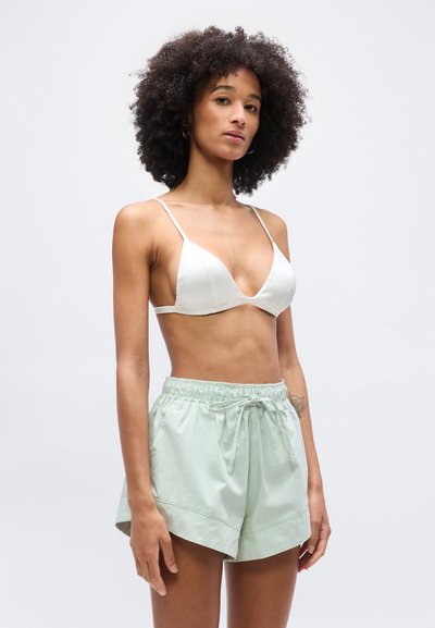 Γυναίκα με σγουρά μαλλιά που φοράει λευκό τρίγωνο σουτιέν και ανοιχτό πράσινο σορτς με κορδόνι, στέκεται μπροστά σε απλό φόντο.