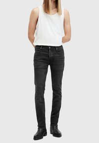 Černé džínové kalhoty slim fit, které mají pět kapes, poutka na opasek a jemné detaily švů. V kombinaci s bílým tílkem bez rukávů.