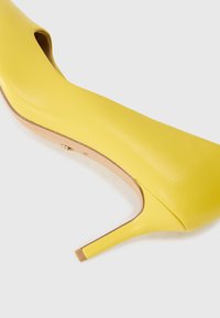 Pump giallo con tacco a stiletto, realizzato in pelle liscia, punta affilata e tacco sottile. Dettaglio di taglio sottile sul lato.
