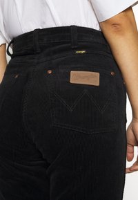 Černé kordové kalhoty s vysokým pasem, vybavené koženou nášivkou Wrangler a kontrastním šitím na zadních kapsách. Jsou přítomny měděné kovové detaily.