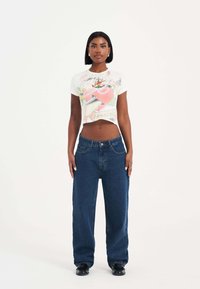 Camiseta gráfica blanca con un diseño colorido, combinada con jeans de mezclilla azul oscuro de corte holgado. El modelo se encuentra de pie contra un fondo sencillo.