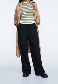 Beige trenchcoat over een groen-wit gestreepte ribbeltop, gecombineerd met zwarte wijde broek en zwarte schoenen met een stevige zool.