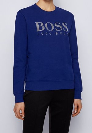 Personne portant un sweat-shirt bleu à manches longues avec « BOSS HUGO BOSS » en clous argentés et un pantalon noir sur un fond clair uni.