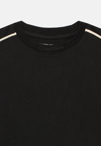 Melnā T-kreklā ar apaļu kakla izgriezumu, ar gludu tekstūru un Calvin Klein logotipu uz apkakles. Balti akcentu svītri uz pleciem.