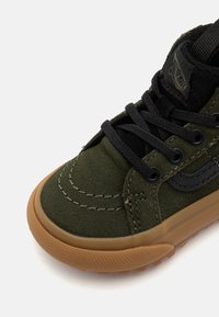 Mörkgrön mockaskor med svarta snören, stickade detaljer och en texturerad gummisula. Har en diskret logotyp på sidan.