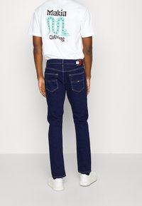 Tmavě modré džíny s klasickým střihem, pěti kapsami a kontrastním zlatým prošitím. Kombinovány s bílým tričkem s barevným grafickým motivem.