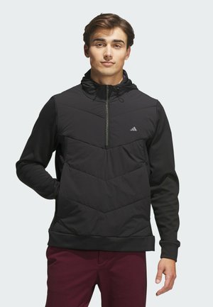 Giovane uomo con un pullover imbottito nero con cappuccio e pantaloni viola, in piedi con una mano in tasca davanti a uno sfondo semplice.