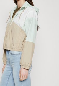 Kurtka typu wiatrówka w beżowych i miętowych odcieniach, z kapturem, zapięciem na zamek, elastycznymi mankietami i luźnym krojem. Gładka tekstura, casualowy design.