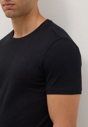 Homme portant un T-shirt noir uni à manches courtes et col rond, debout contre un mur de couleur claire, légèrement tourné vers la gauche.