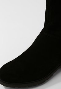 Musta mokkanahkainen nilkkaboots, jossa on pyöreä varvastila, teksturoitu pinta ja litteä kumipohja. Sivusaumassa on näkyvää ompelua.