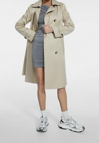 Gabardina beige con un diseño de doble botonadura y hombreras, vestida sobre un vestido gris ajustado y combinada con zapatillas plateadas.