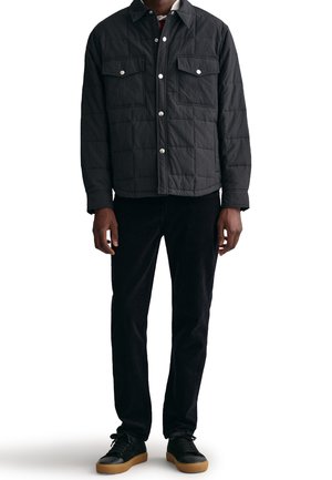 Mężczyzna ubrany w czarną pikowaną kurtkę zapinaną na guziki z klapkowymi kieszeniami, czarne spodnie i czarne trampki z gumowymi podeszwami, stojący na białym tle.