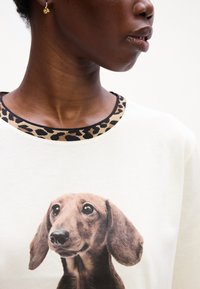 Biele tričko s grafikou hlavy hnedého dalmatína. Výstrih má lemovanie s leopardím vzorom. Hladká textúra látky.