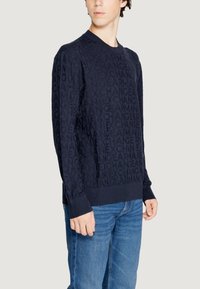 Felpa blu navy con motivo logo in rilievo su tutta la superficie, polsini a costine e colletto girocollo, abbinata a jeans blu chiaro.