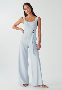 Combinaison bleu clair avec encolure carrée, jambes larges, ceinture avec détail de boucle, et texture lisse. Modèle posant avec une main sur la hanche.