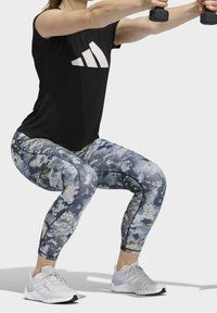 Zwarte sportshirt met wit logo, leggings met een gekleurd patroon en lichtgrijze schoenen. Onderwerp voert een squat uit met dumbbells.