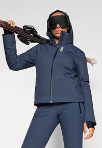 Námořnická sněhová bunda a kalhoty v sadě, vyrobené z voděodolného materiálu, s kapucí, zipovým uzávěrem a elegantním designem. Lyže drženy na rameni, velké brýle na lyže nasazeny.