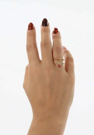Bague en or avec une bande texturée, ornée d'un petit pendentif avec des ailes et un accent rouge, portée sur une main aux ongles marron et décorés.