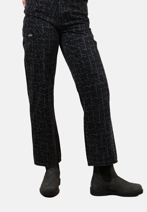 Pantalon noir avec un motif floral subtil porté avec des bottines noires sur une personne debout devant un fond blanc.