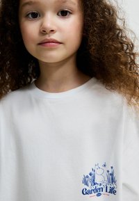 Vit bomull t-shirt med blåtryckt design av en tecknad karaktär omgiven av blommor, med texten "Garden Life" på framsidan.