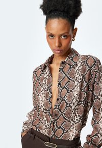 Blusa con stampa in rettile marrone e nera, con bottoni frontali, maniche lunghe e colletto, abbinata a pantaloni marroni a vita alta.