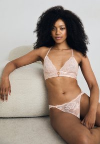 Ensemble bralette et string en dentelle rose pâle, présentant des bords festonnés, des bretelles réglables et un détail de fermeture central sur le soutien-gorge.