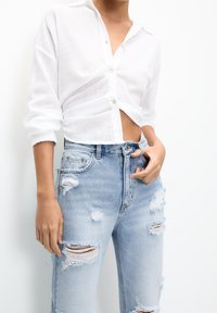 Chemise en lin blanche à manches longues et boutons, associée à un jean en denim bleu clair déchiré avec une coupe légèrement ample.