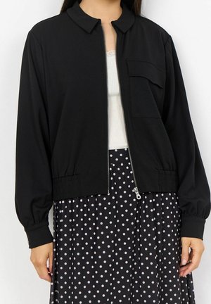 Kobieta ubrana w czarną kurtkę na zamek błyskawiczny na białym topie z koronkową lamówką i czarną spódnicę w białe kropki, stoi z rozluźnionymi ramionami.
