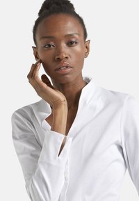 Camicia bianca con colletto, realizzata in tessuto morbido, dotata di pattina e dettagli unici ai polsini, indossata da un modello con uno sfondo neutro.