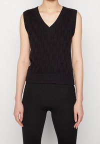 Zwart mouwloos kabelgebreid sweatervest met V-hals, gedragen over nauwsluitende zwarte broek, gedragen door een staand persoon tegen een effen achtergrond.