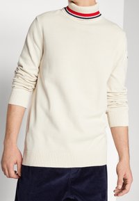 Beiger Rollkragenpullover mit Rippkragen, der rote und marineblaue Streifen aufweist. Weiches Strickmaterial, lockere Passform, gerippte Bündchen und Saum.