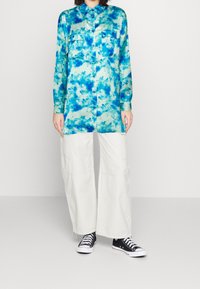 Camicia a maniche lunghe con bottoni in stampa tie-dye blu e aqua, abbinata a pantaloni bianchi a gamba larga e sneakers nere, in piedi su uno sfondo semplice.