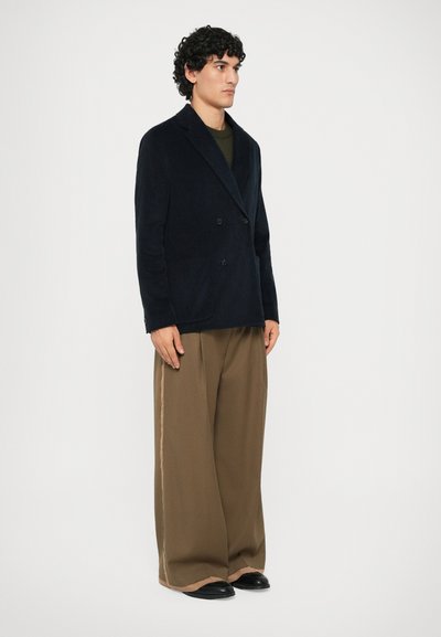 Sacou bleumarin cu două rânduri de nasturi, dintr-un material texturat, asortat cu pantaloni largi maro, având o dublă dungă contrastantă pe lateral și pantofi negri.