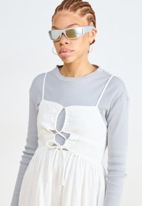 Vestido blanco con detalles de corte y lazos, superpuesto a una camiseta de manga larga de canalé gris claro. La modelo lleva gafas de sol grises reflectantes.