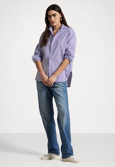Gestreifte Hemdbluse in Lavendel und Weiß, mit lockerer Passform und hochgekrempelten Ärmeln, kombiniert mit hellblauen Jeans und beigen Sneakers.