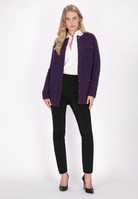 Fialový pletený cardigan s knoflíkovým zapínáním, nošený přes bílou košili, v kombinaci s černými úzkými kalhotami a černými lodičkami. Hladká textura.