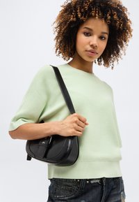 Νεαρή γυναίκα με σγουρά μαλλιά που φοράει ένα ανοιχτό πράσινο πουλόβερ με κοντά μανίκια και κρατάει μια μαύρη δερμάτινη τσάντα ώμου με λεπτομέρειες αγκράφας.