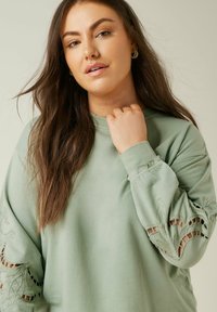 Sudadera verde claro con detalles recortados bordados en las mangas, de manga larga y cuello redondo, fabricada con un material suave y texturizado.