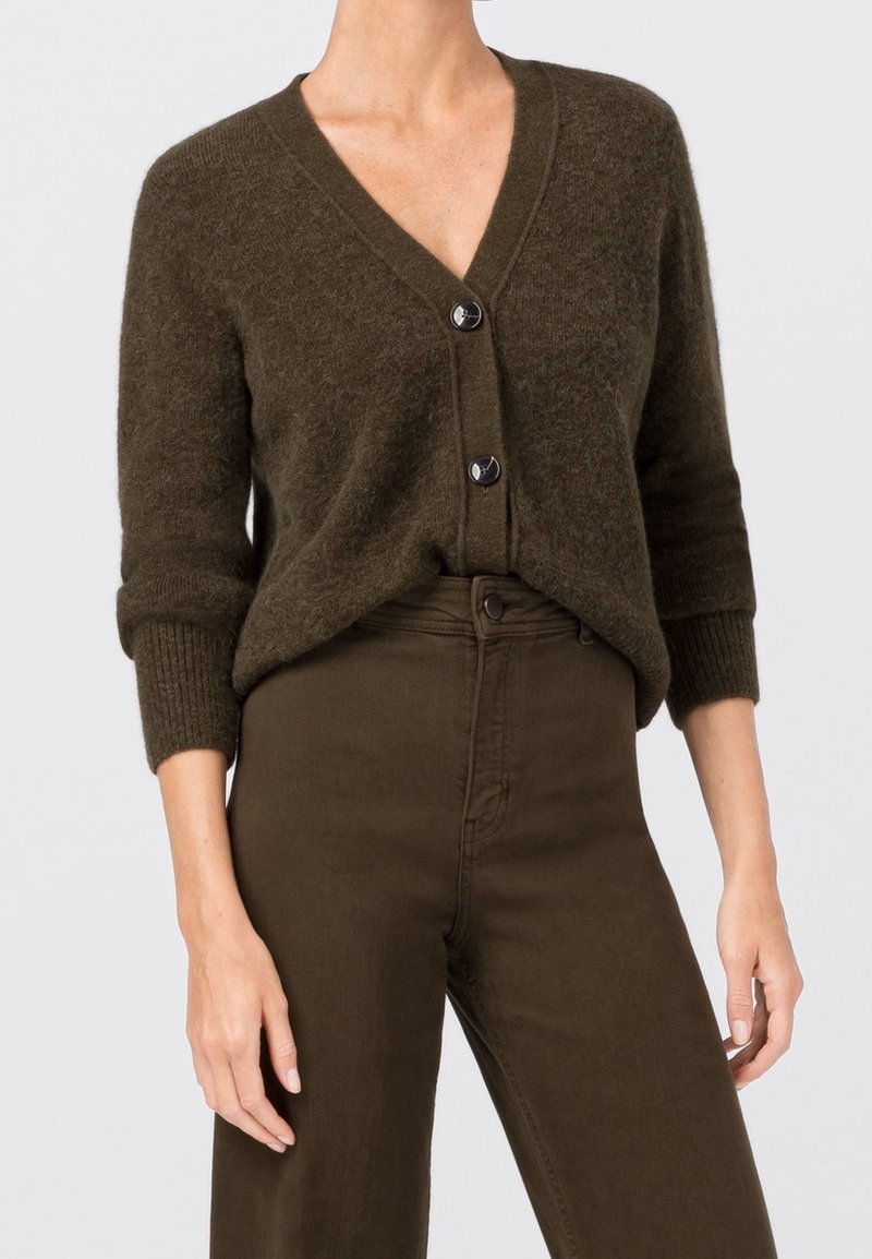 Femme portant un cardigan duveteux à col en V couleur olive foncé avec deux boutons et un pantalon assorti taille haute, debout devant un fond uni.