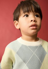 Pull léger bleu avec des accents crème présentant un motif en losanges. Le modèle a les cheveux courts et foncés devant un fond rouge.